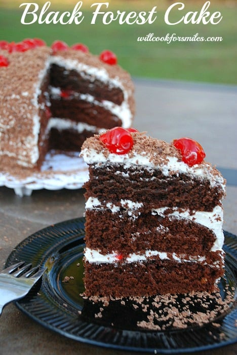 Black Forest Cake slice on a black plate with a fork to the left and the rest of the cake in the background
