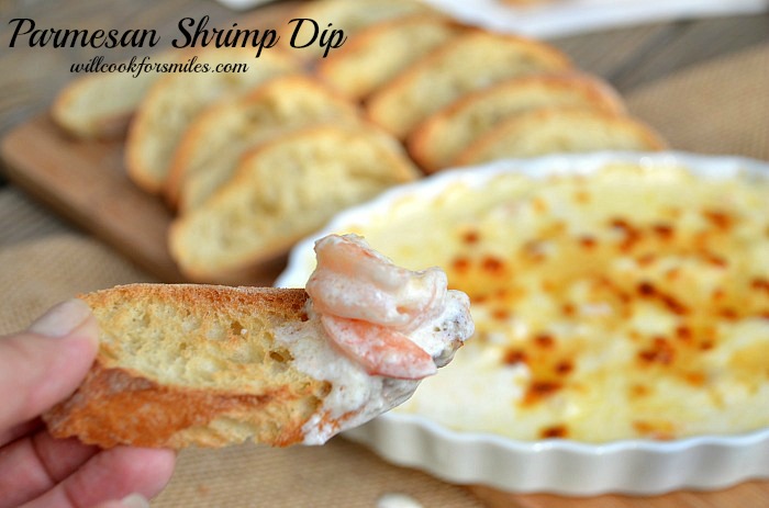 holding a piece of crusty bread with Parmesan shrimp dip on it and the rest in a casserole dish with crusty bread sliced next to it on a cutting board