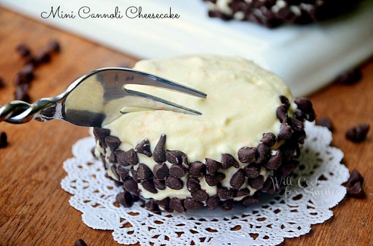 fork cutting into cannoli cheesecake on decorative white napkin with white rectangular plate and additional cannolis in background all on wooden table