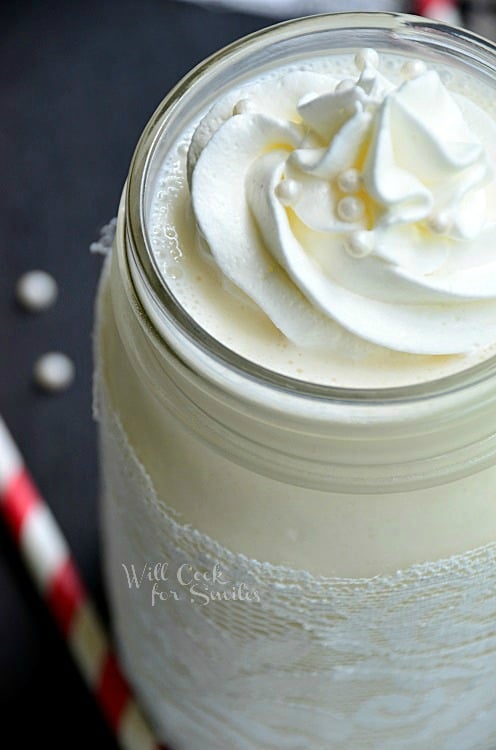 close up picture of mason jar filled with white wedding cake milkshake on a black table with red and white striped straws laying around jar