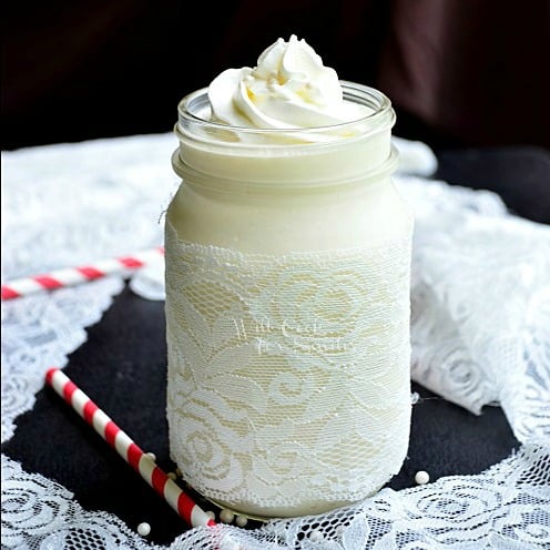 White Wedding Cake Milkshake in a mason jar with lace around the bottom of the mason jar and whipped cream and candy pearls on top with a red and white straws next to it
