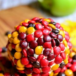 dessert cheese ball covered in M&Ms on wood cutting board with apple in background to the left