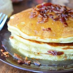 stack of maple bacon pancakes on black plate with fork to the left and syrup jar in background to the left