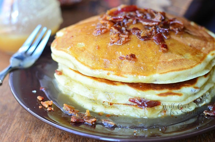 stack of maple bacon pancakes on black plate with fork to the left and syrup jar in background to the left