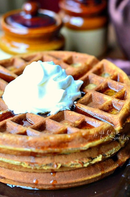 stack of gingerbread waffles with a whipped topping on small plate with fork at the bottom right of plate on a brown placemat. 2 brown and tan jars in background to the left