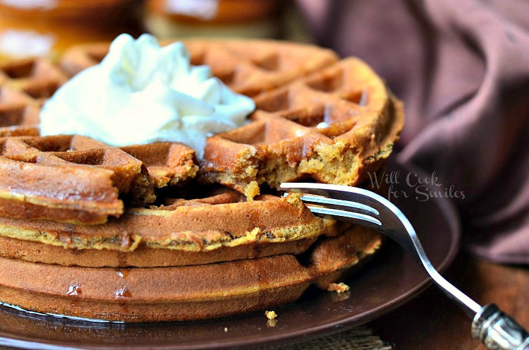 close up horizonal ginger bread waffles with whipped cream on top on a brown plate with a fork to the right