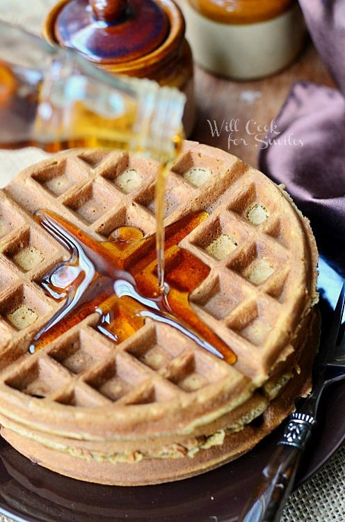 pouring syrup over ginger bread waffles with whipped cream on top on a brown plate with a fork to the right