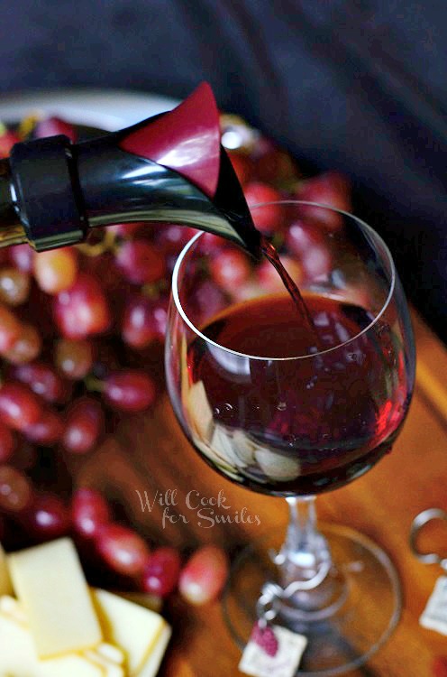 pouring red wine into a wine glass that is sitting on a cutting board with cheese and grapes