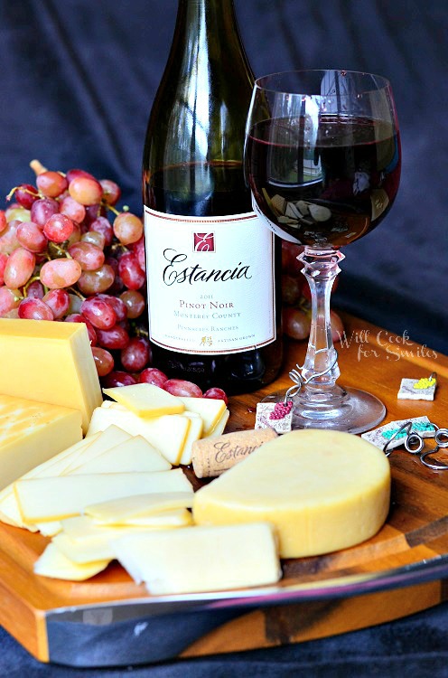 glass of red wine on cutting board with grapes and several different types of cheese and bottle of wine next to cheese
