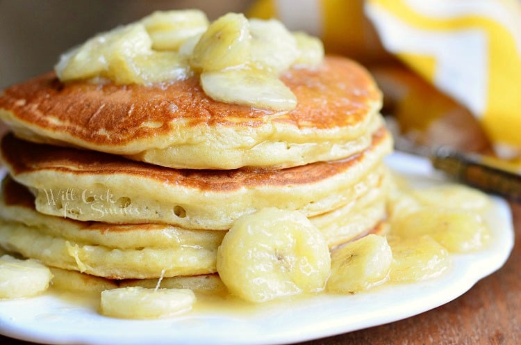 stack of bananas foster pancakes on white plate on wood table with yellow and white cloth in background