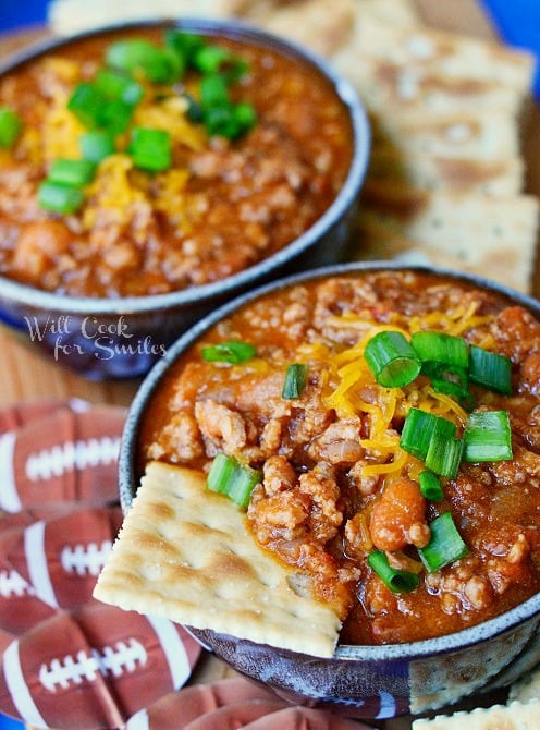 above view of 2 small bowls of boozy chili on a blue platter with cracker scattered around bowls. Chili topped with shredded cheese and chives