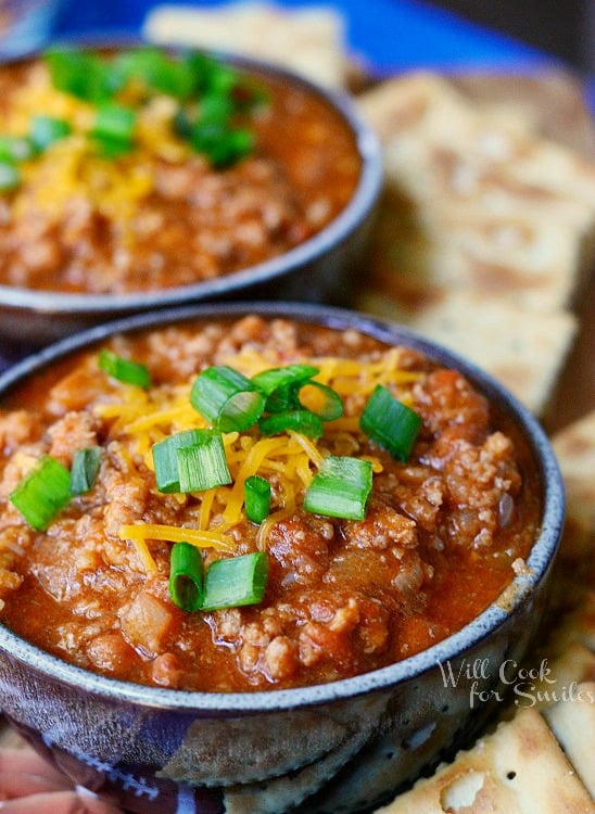 2 small bowls of boozy chili on a blue platter with cracker scattered around bowls. Chili topped with shredded cheese and chives