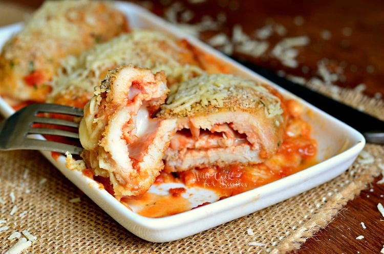 rectangular plate with chicken parmesan roll ups and fork holding one bite above first rollup. Plate is on a brown placemat on a wood table.