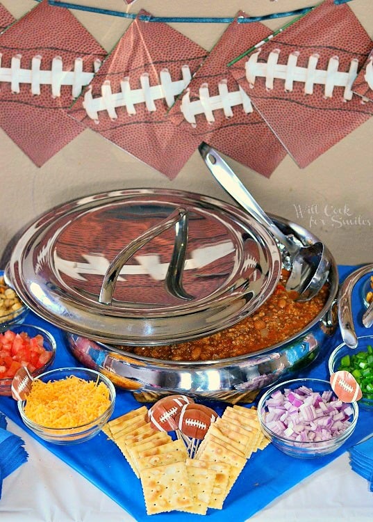 large stainless steel bowl filled with chili topped with lid and silver spoon on a football themed dressed table with cracker and toppings for chili in front of silver bowl