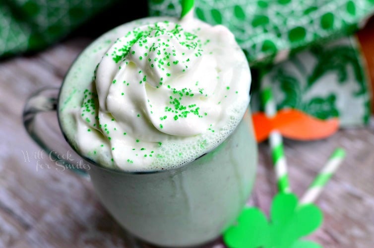 clear glass coffee mug filled with st patrick's day skinny milkshake creamy mint chocolate chip on a wood table with st patties day decore scattered on table