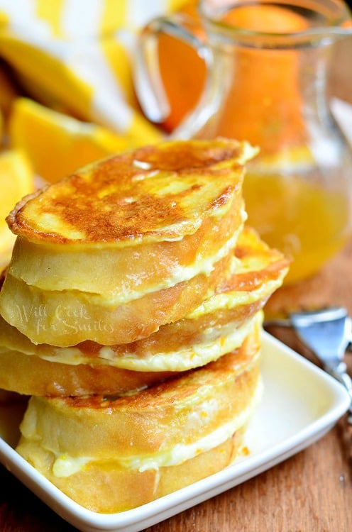 stack of orange creamsicle french toast slices on a white plate on wooden table with yellow and white cloth in background with syrup jar
