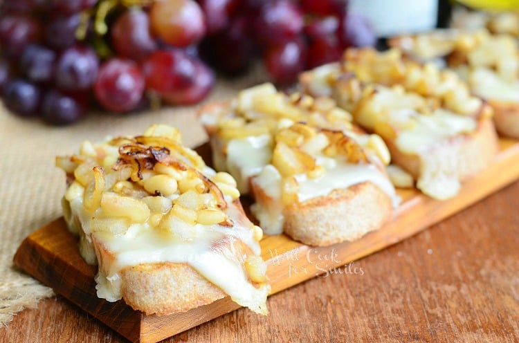 several Presidents cheese crostinis lined up on a wood plank on wood table with grapes, bottle of wine and cheese package in background