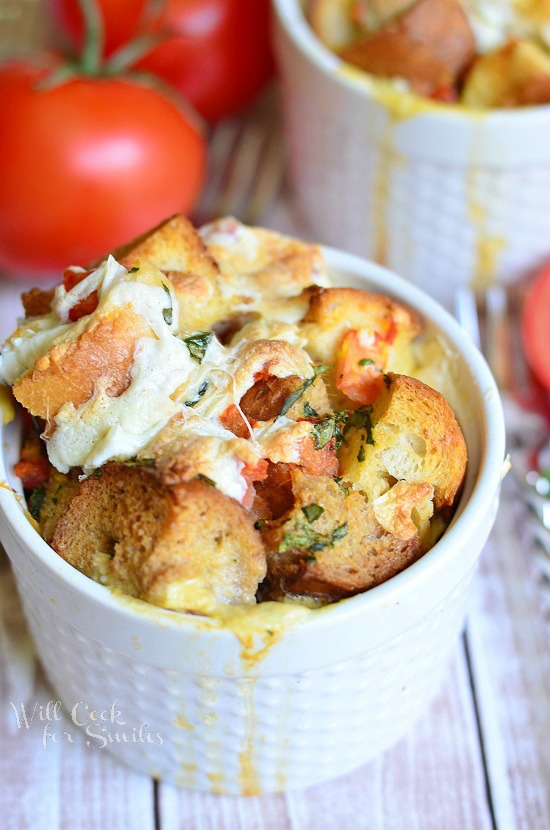 small white crock filled with tomato basil bread pudding on a white table with fork to the right of crock and tomato and additional bread pudding in background