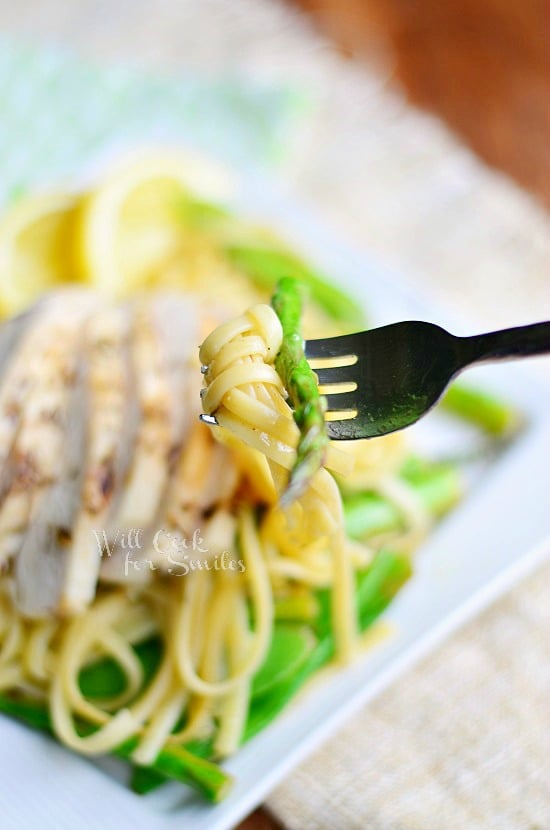 Linguine with asparagus on a fork with linguine, Chicken, Asparagus and Snow Peas on a white square plate and with slices of lemon and a fork