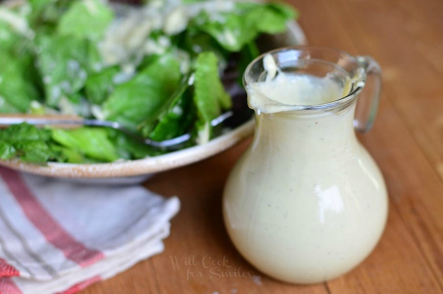 small dressing jar filled with light caesar dressing on a wood table in fron of a ceramic bowl filled with caesar salad