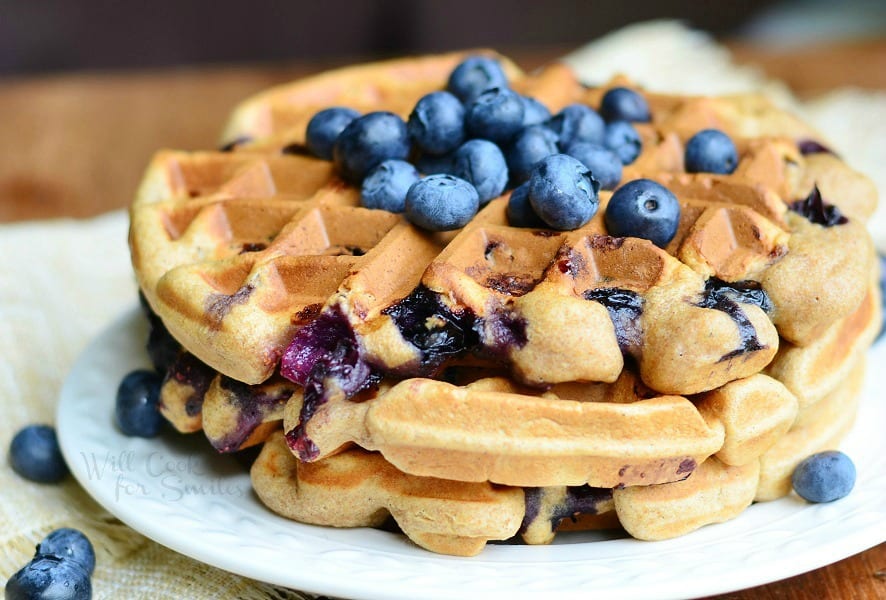 close up view of round white plate with blueberry waffles and blueberries topping the waffles all on a wood table with tan cloth and blueberries below plate