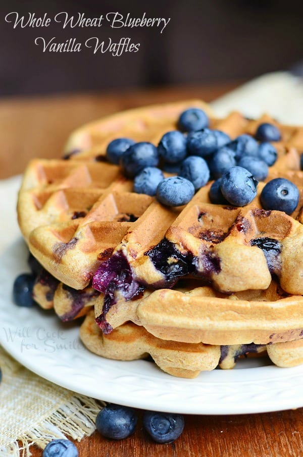 round white plate with blueberry waffles and blueberries topping the waffles all on a wood table with tan cloth and blueberries below plate