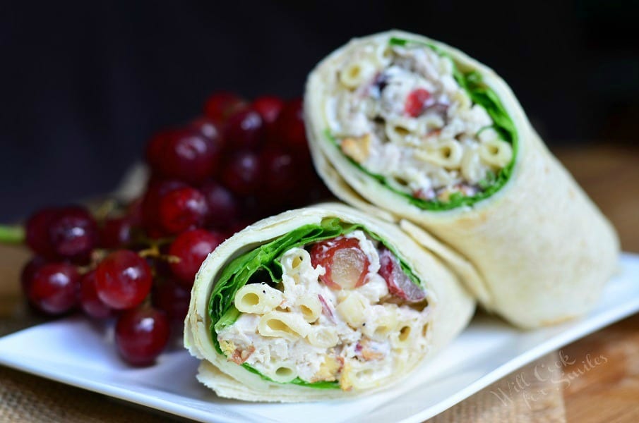 A wrap made with chicken deluxe salad and lettuce on a plate with a grapes in background.