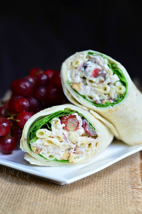 A wrap made with chicken salad cut in half and on a white plate with a bunch of grapes.