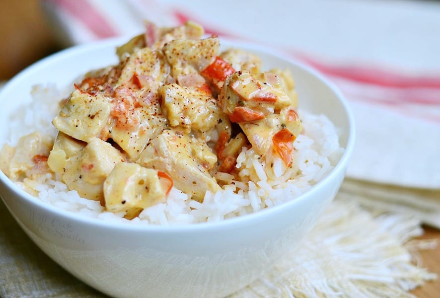 close up view of white bowl filled to the brim with creamy cajun chicken rice on a white table cloth on wooden table with another white and red cloth in background