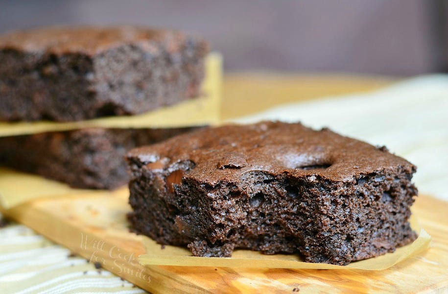 close up view of 2 dark chocolate chocolate chip brownies stacked with wax paper in between each stacked in background with 1 brownie in foreground on a wooden cutting board on a tand and white placemat on wood table