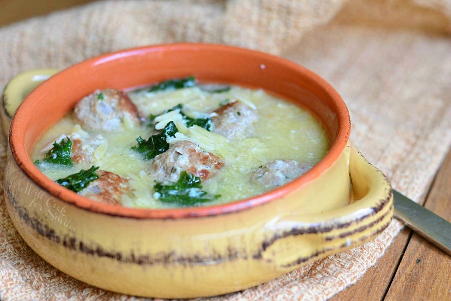 close up view of Orange and tan clay crock with Italian Wedding Soup with turkey meatballs and Orzo sitting on a tan table cloth on a wood table