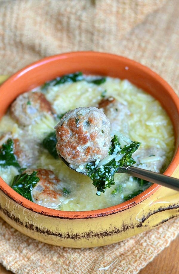 above view of Orange and tan clay crock with Italian Wedding Soup with turkey meatballs and Orzo sitting on a tan table cloth on a wood table with a spoon picking up a meatball