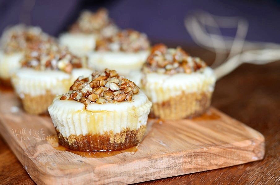 close up view of 6 pecan mini cheesecakes lined up in 2 rows on a wood cutting board with maple syrup drizzled across the tops of the cupcakes