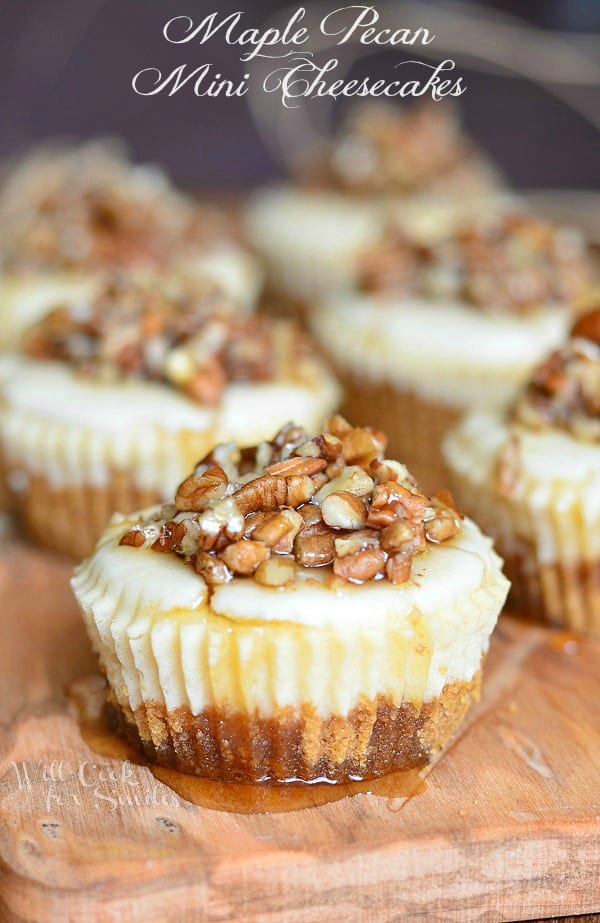 One cheesecake in foreground with 6 pecan mini cheesecakes lined up in 2 rows on a wood cutting board with maple syrup drizzled across the tops of the cupcakes