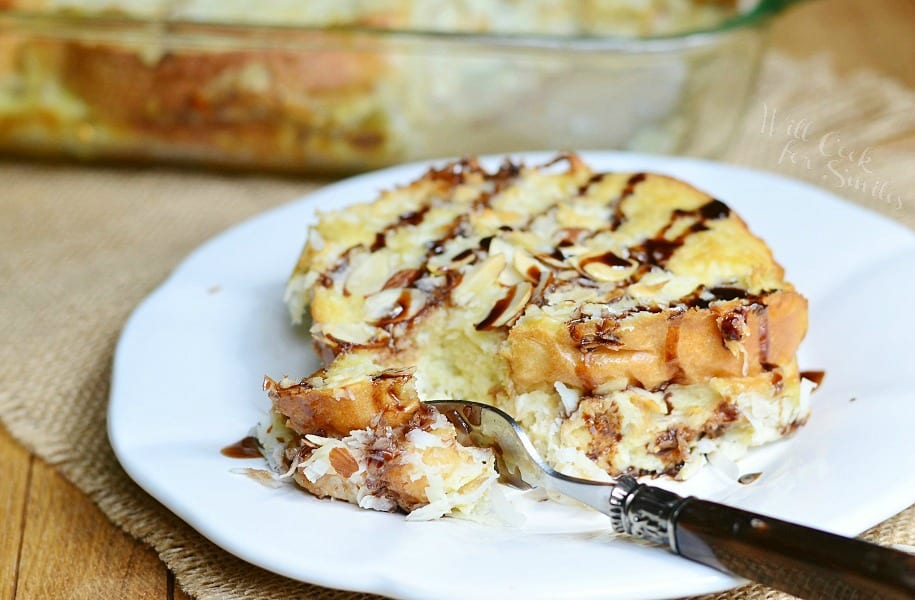 close up view of one portion of almond joy french toast bake on a round white plate on tan placemat with baking dish and rest of bake in background and fork taking piece of french toast