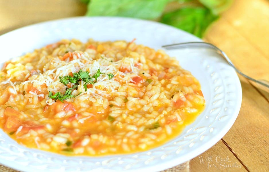 close up veiw of a white decorative rimmed bowl filled with creamy tomato basil risotto on a tan placemat on wooden table with leaves of basil as a garnish
