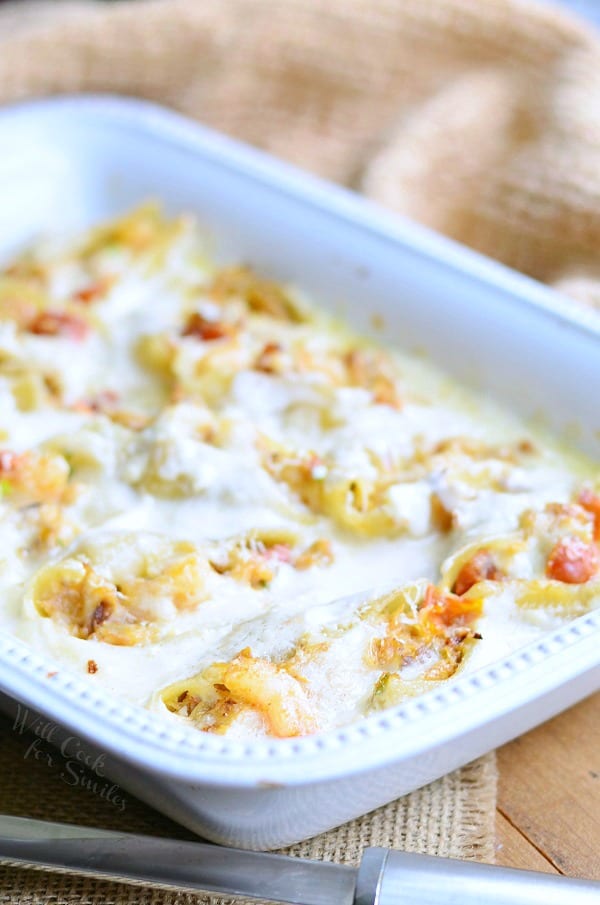 white baking dish filled with seafood alfredo stuffed shells on a tan placemat on wooden table with a silver serving spoon at bottom left of baking dish