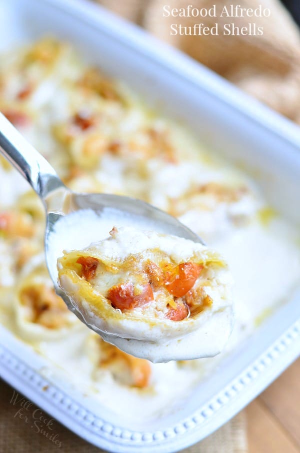 white baking dish filled with seafood alfredo stuffed shells on a tan placemat on wooden table with a silver serving spoon at bottom left of baking dish with a spoon holding one bite above the baking dish