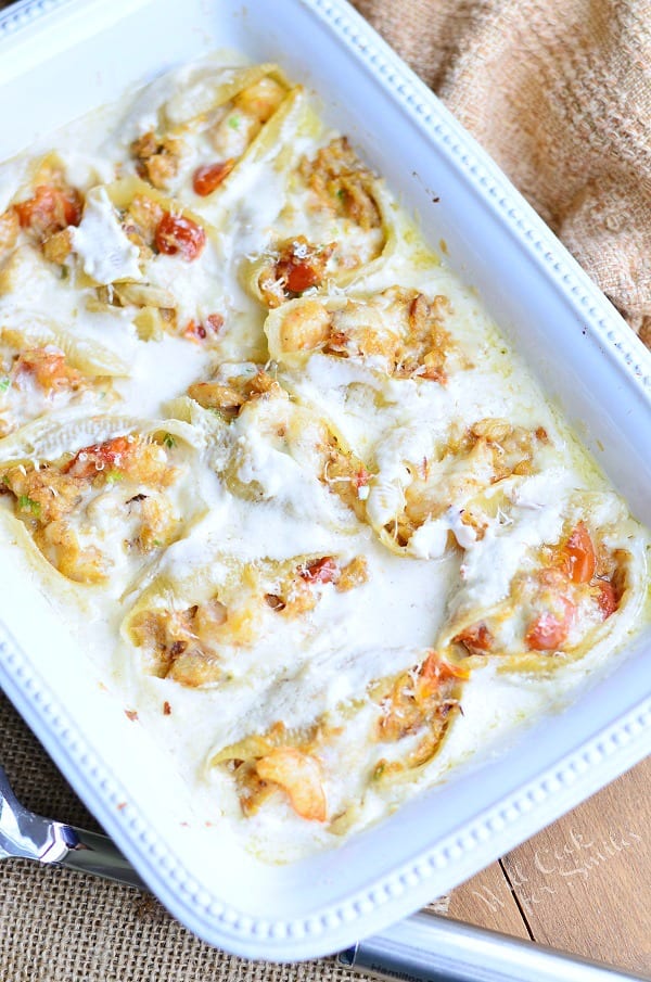 white baking dish filled with seafood alfredo stuffed shells on a tan placemat on wooden table with a silver serving spoon at bottom left of baking dish