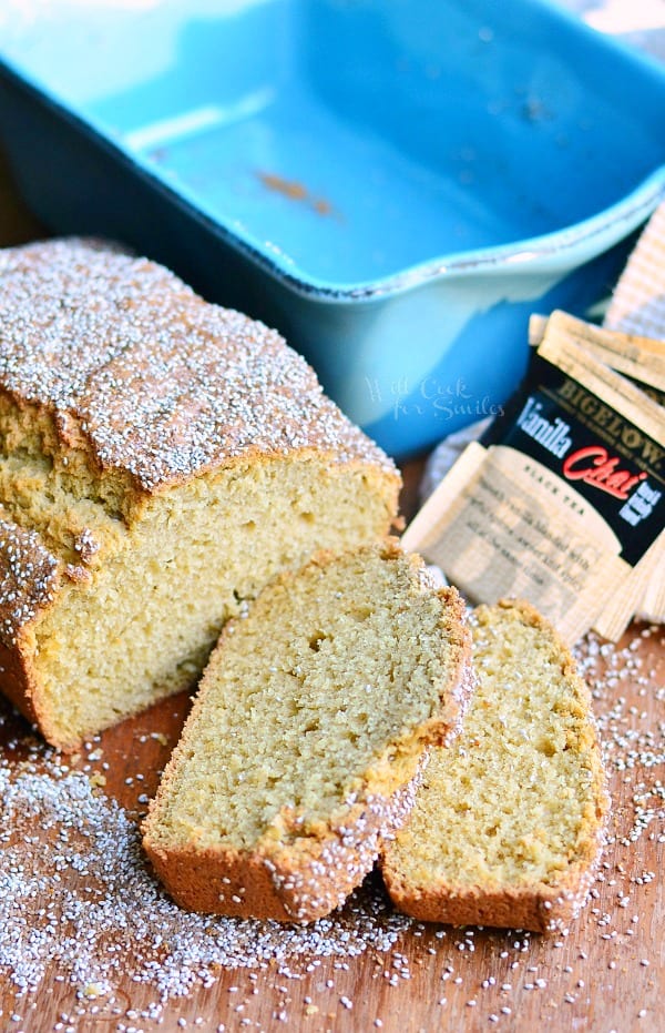2 slices of vanilla chai tea bread cut from loaf laying on a wooden table with the rest of loaf behind and a blue baking dish in background with tea bags laying on tan cloth