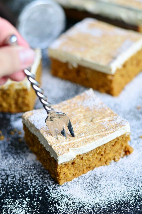 fork being held in cake square in foreground of 3 squares of gingerbread cake on a black table dusted with powdered sugar and a baking dish with more cake in the background