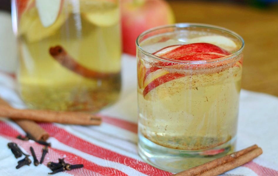 close up view of small glass filled with spiced apple sangria with sliced apples floting near the top of the glass while the glass is on a white and red cloth and a cinnamon stick rests at bottom of glass. A glass pitcher rests in background filled with spiced apple sangria and an apple to it's right.