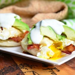 close up view of white rectangular plate with an avocado bacon ranch breakfast on a wooden table with brown cloth topped with a white and green cloth