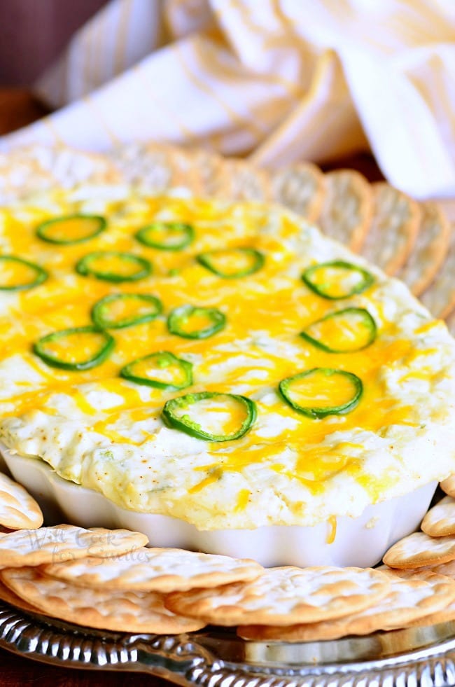 decorative white bowl filled with cheesy jalapeno dip centered in between crackers laying on a silver platter with a white and gold cloth in the background