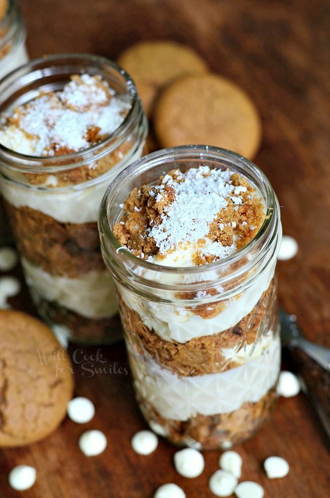 3 mason jars filled with no bake white chocolate cheesecake gingerbread trifles on a wooden table with white candy scattered on table