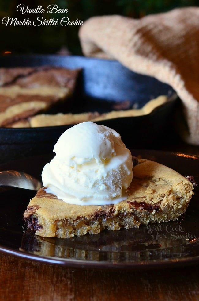 1 slice of vanilla bean marble skillet cookie on a black plate topped with a scoop of ice cream on a wooden table. The skillet with the rest of the cookie remains in the background with a tan cloth draped over the side.