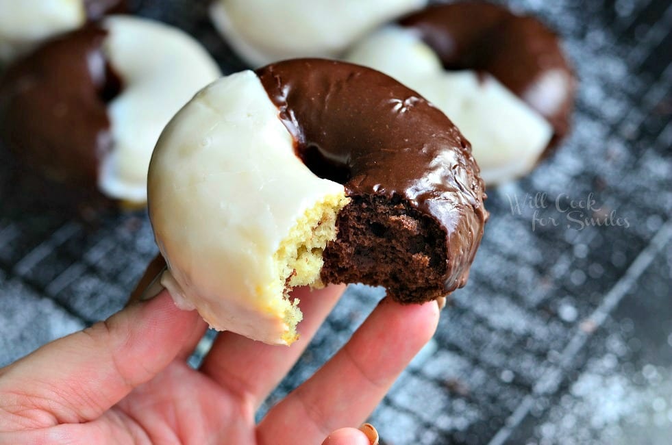 hand holding 1 donut with a bite taken out above Black and white glazed donuts on a cooling rack on a black table with powdered sugar on table