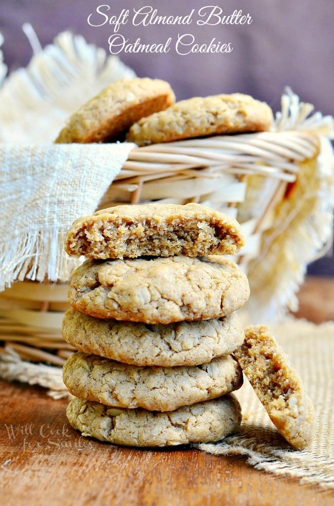Soft Almond Butter Oatmeal Cookies stacked up on a table with a basket in the background with cookies in it with the top one cut in half