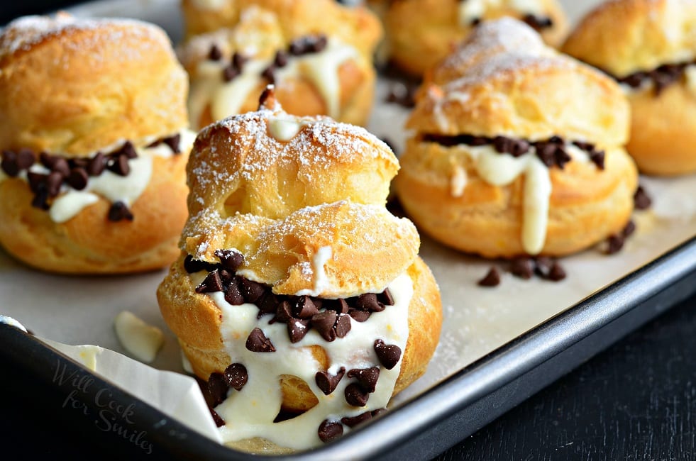 close up view of a Baking pan with wax paper holding cannoli pastry cream puffs on a black table