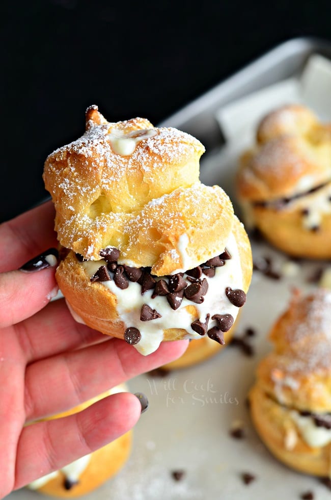 hand holding on cannoli above a Baking pan with wax paper holding cannoli pastry cream puffs as viewed from above on a black table